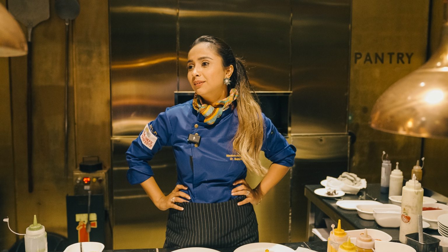 Sakirat Waraich in a blue chef coat inside a professional kitchen, NILA-branded jacket visible.